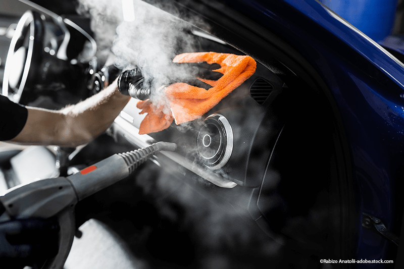 Eine Person reinigt das Armaturenbrett eines blauen Autos mit Dampf und einem orangefarbenen Tuch; Innenaufnahme.