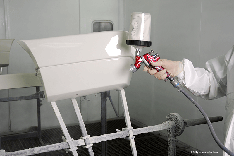 Eine Person besprüht einen Gegenstand in einer Lackierkabine mit weißer Farbe mithilfe einer Spritzpistole.