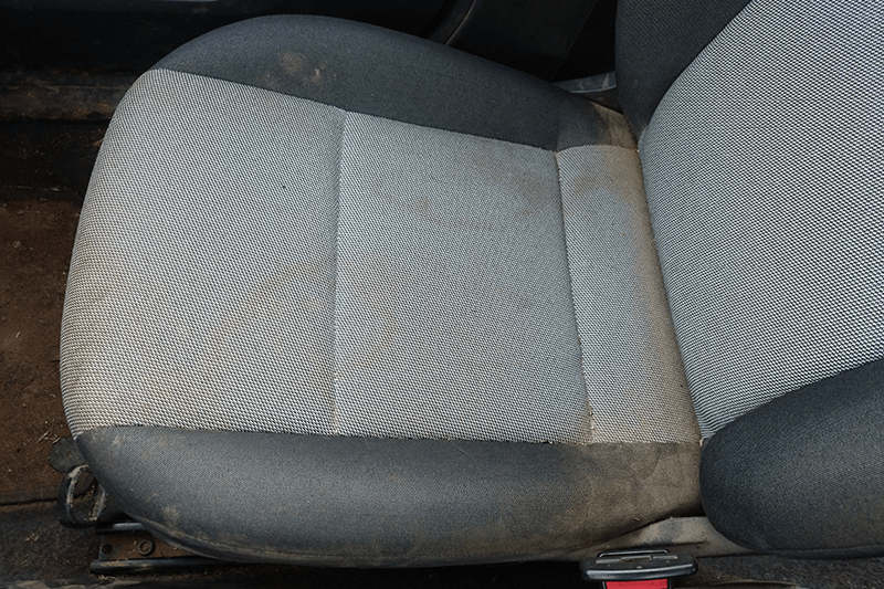 Schmutziger Autositz mit hellgrauem Stoff und dunkelgrauen Kanten.
