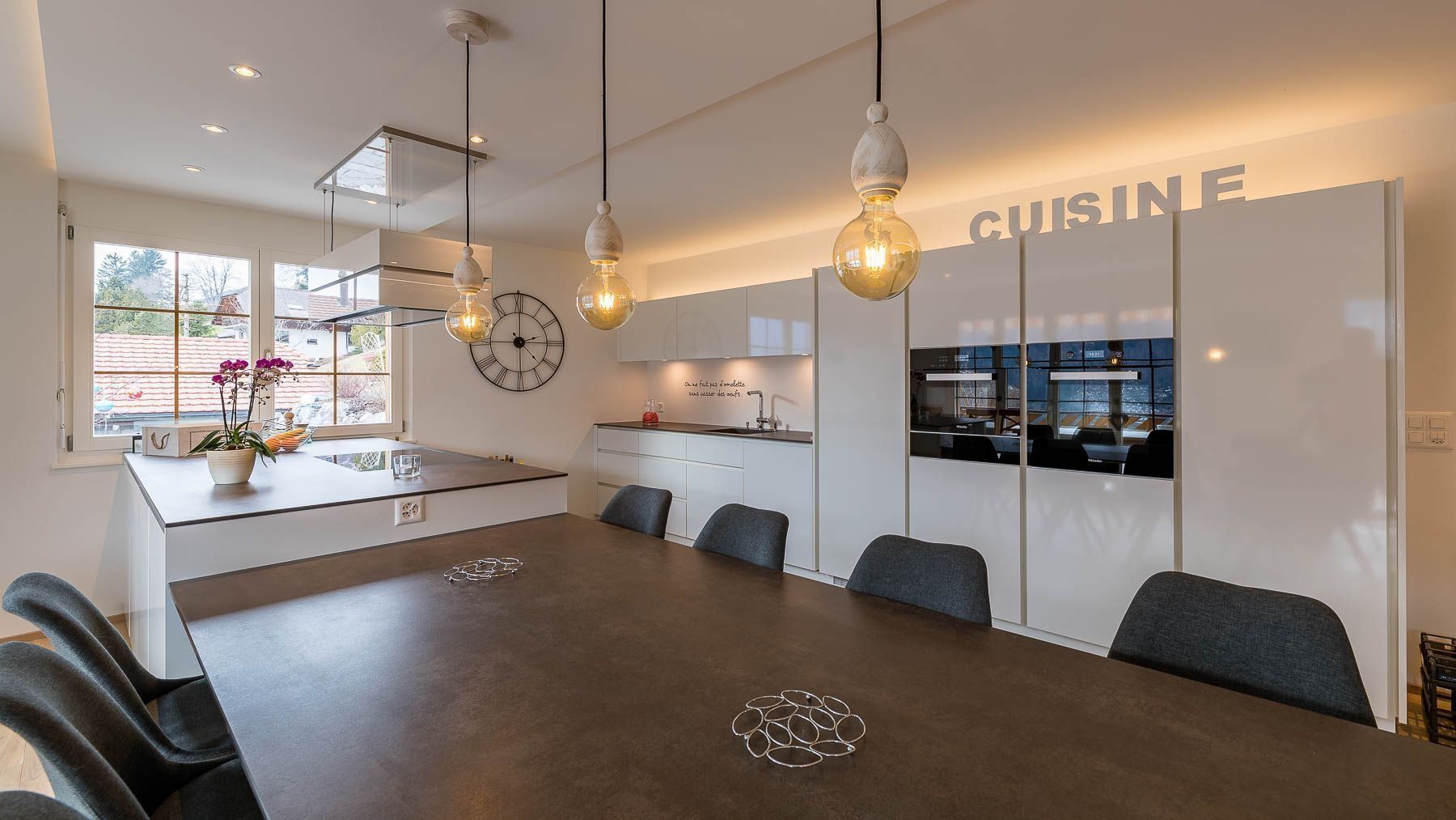 Cuisine moderne avec armoires blanches, table en bois foncé, luminaires suspendus et panneau lumineux « CUISINE ».