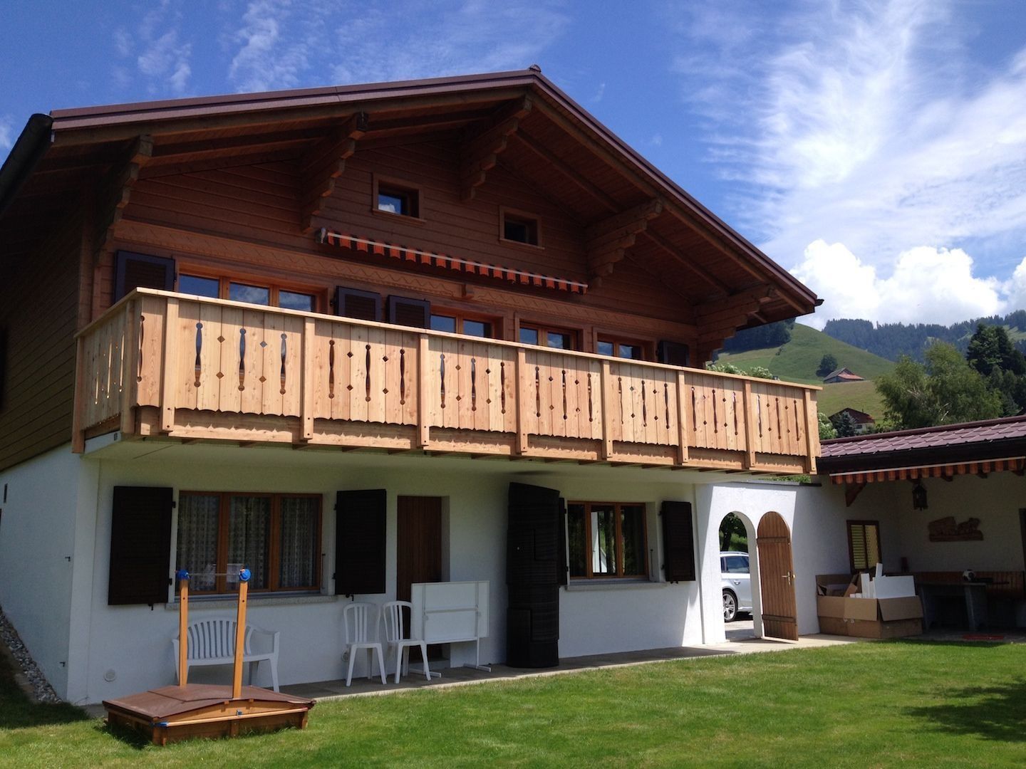 Chalet en bois avec balcon, niveau inférieur blanc, pelouse verte, montagnes en arrière-plan, journée ensoleillée.