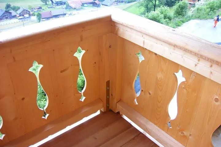 Rambarde de balcon en bois ornée de découpes décoratives, se détachant sur un fond vert flou de bâtiments et de végétation.