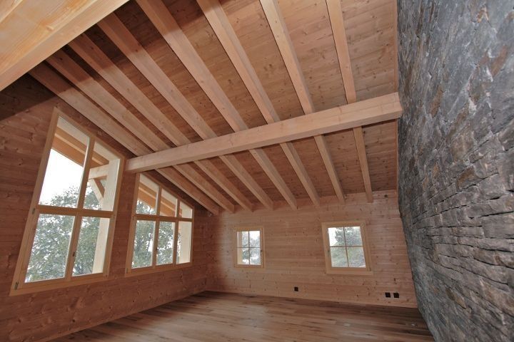 Intérieur d'une pièce avec poutres apparentes au plafond, fenêtres et mur en pierre.