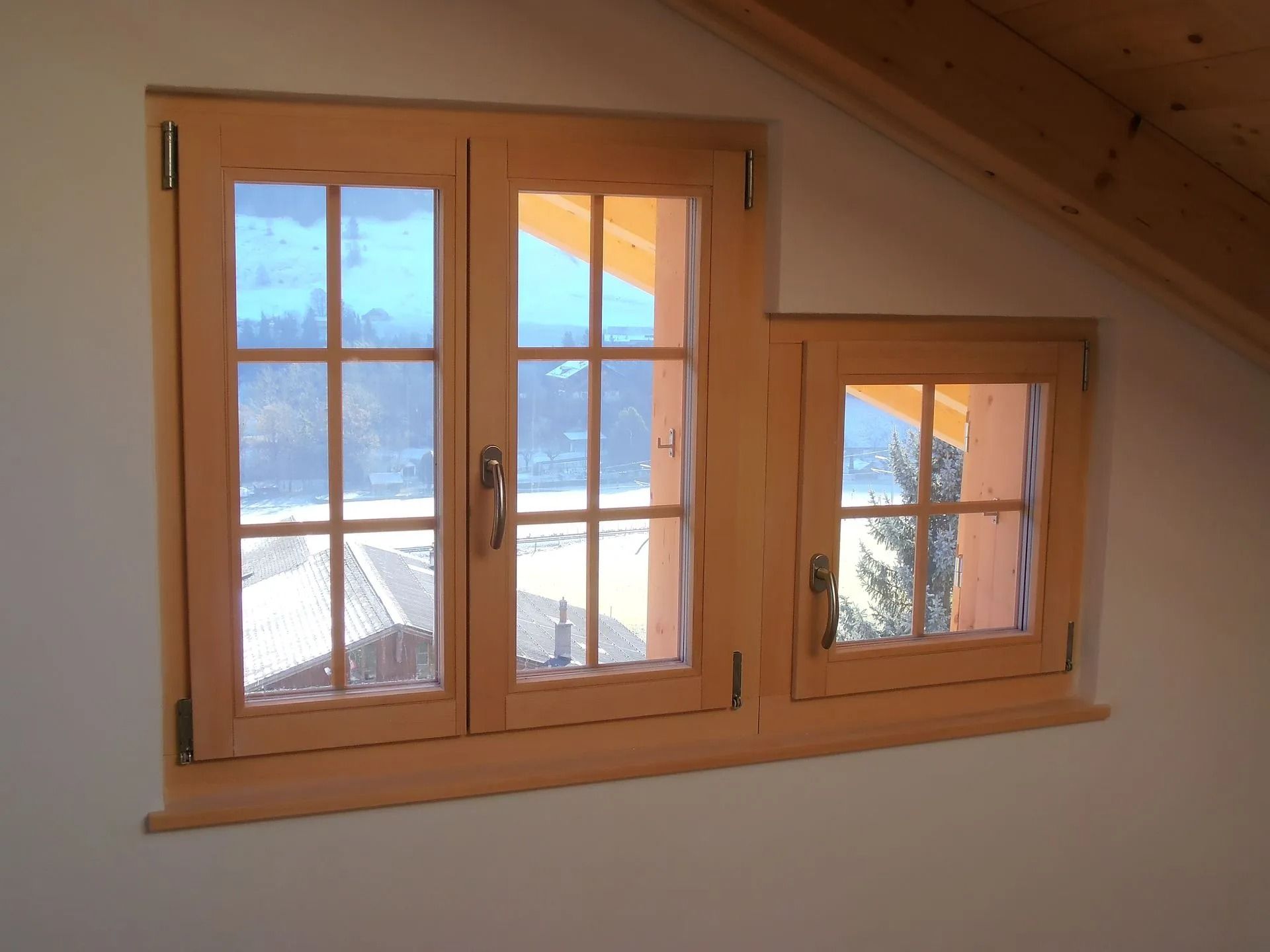 Fenêtres en bois dans une pièce blanche, encastrées dans un mur incliné, avec vue sur un paysage enneigé.