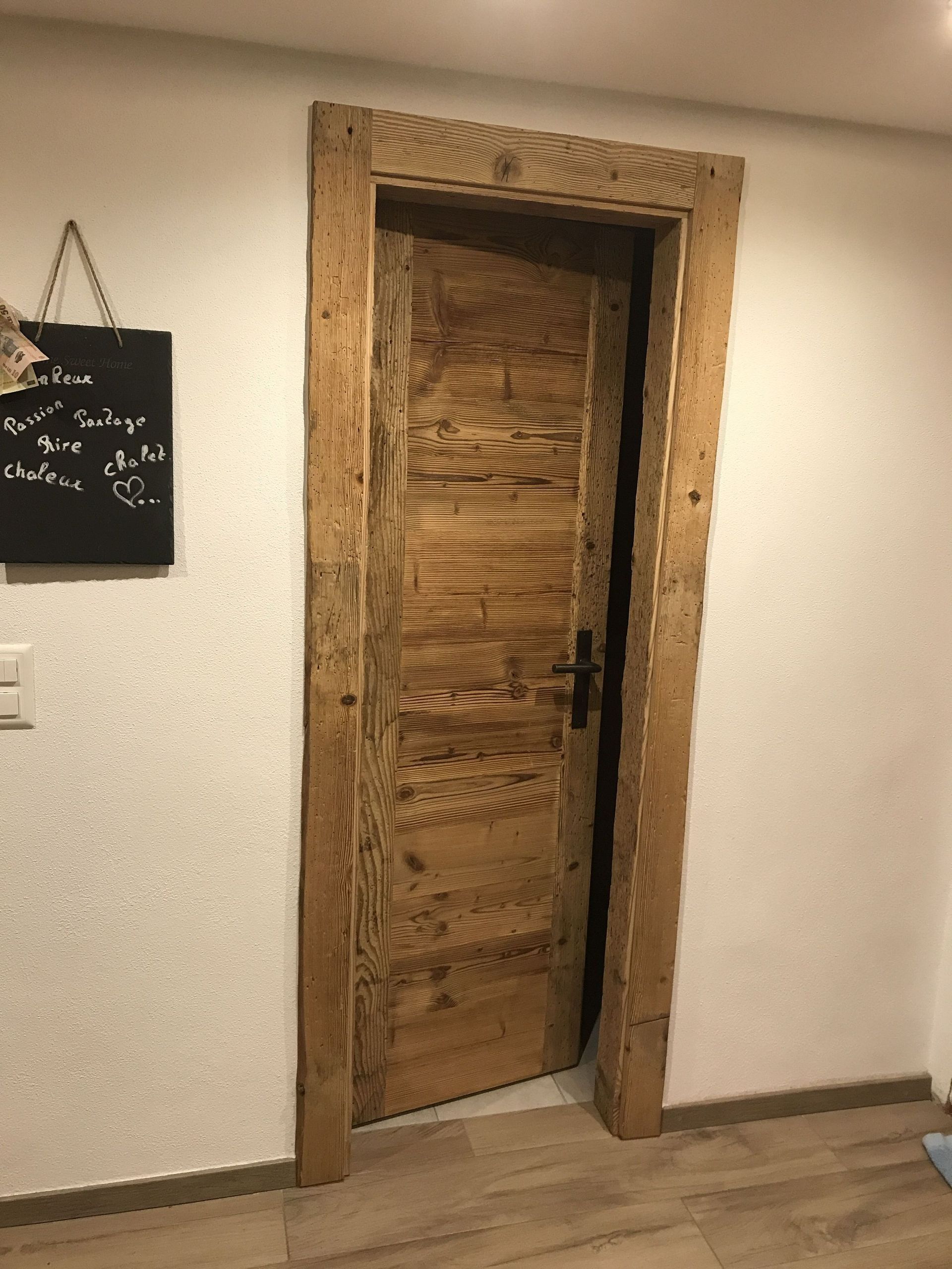 Porte en bois avec détails et cadre sculptés ; sur un mur blanc, un tableau noir à côté.