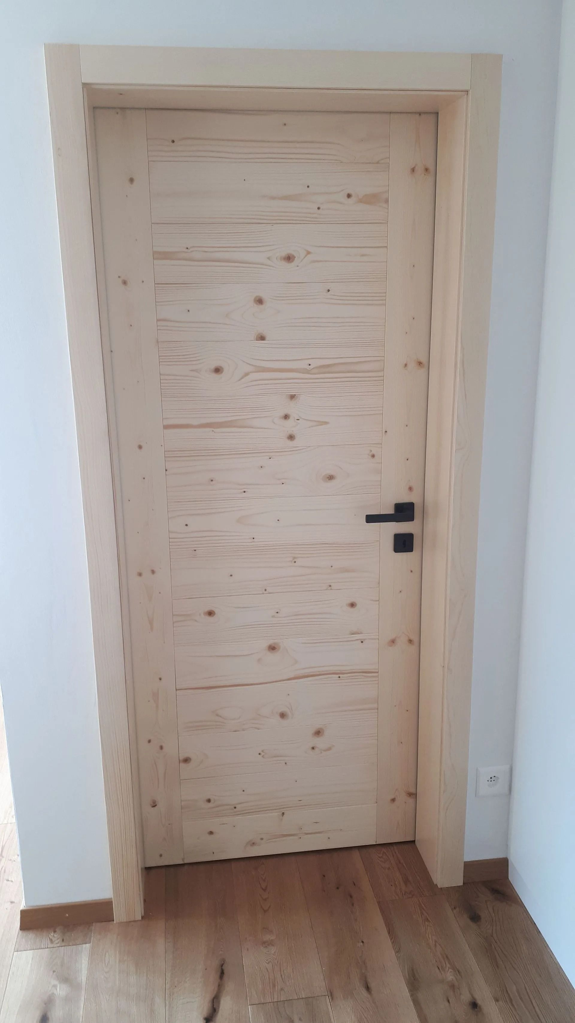 Porte en bois à cadre clair et poignée noire. La porte est encastrée dans un mur blanc au sol en bois.