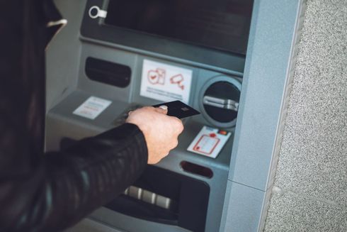 Una mano en la manga de una chaqueta negra sostiene una tarjeta cerca de un lector sin contacto en un cajero automático gris.
