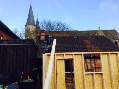 Bauarbeiter auf dem Dach, im Vordergrund Gebäude mit Holzpaneelen, im Hintergrund Kirchturm.