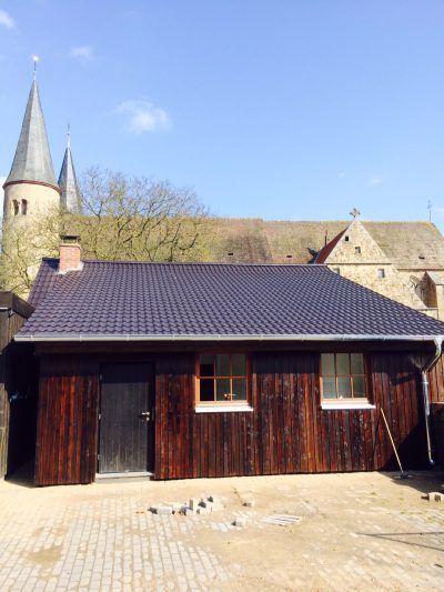 Holzgebäude mit dunklem Dach, Tür und Fenstern; Kirchturm im Hintergrund.