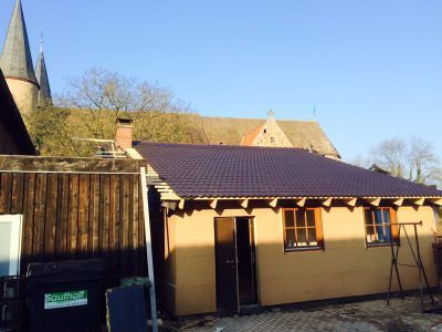 Kleines braunes Gebäude mit dunkelviolettem Dach, braunen Zierleisten und einer dunklen Tür. Im Hintergrund ist eine Kirche.