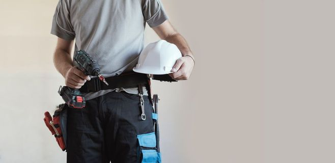 Ein Bauarbeiter hält eine Bohrmaschine und einen Helm in der Hand, trägt einen Werkzeuggürtel und steht vor einem hellen Hintergrund.