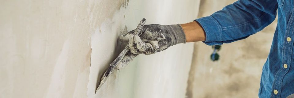 Eine Person mit blauem Hemd und Handschuhen trägt mit einer Kelle eine cremefarbene Substanz auf eine Wand auf.