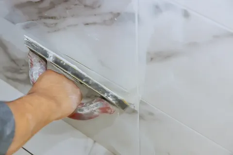 Person, die mit einer Kelle Fugenmörtel zwischen weißen Marmorfliesen aufträgt.