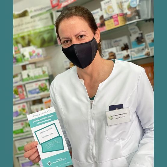 Una persona con bata blanca y mascarilla negra sostiene una caja de producto de color turquesa en una tienda.