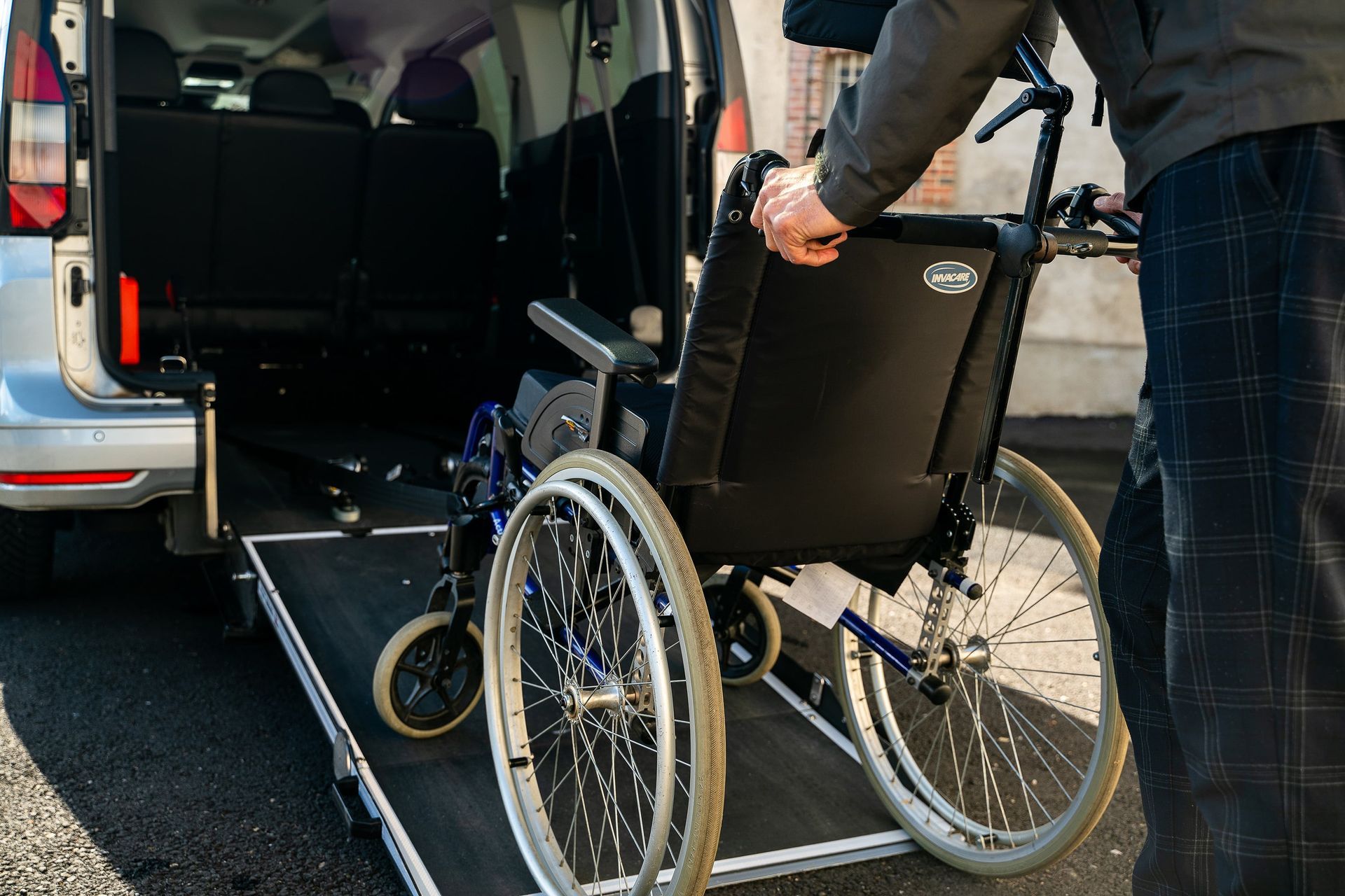 Une personne charge un fauteuil roulant sur la rampe d'une camionnette. Camionnette argentée, fauteuil roulant bleu, cadre extérieur ensoleillé.