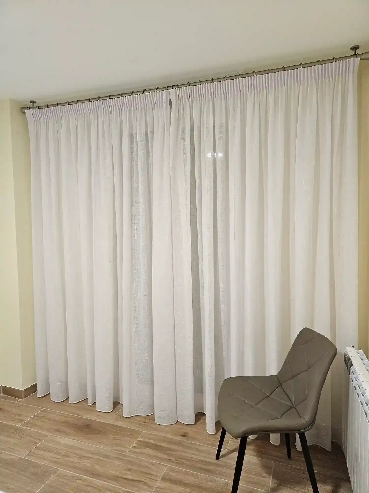 Unas cortinas blancas cubren una gran ventana en una habitación minimalista con una silla gris sobre suelo de madera.
