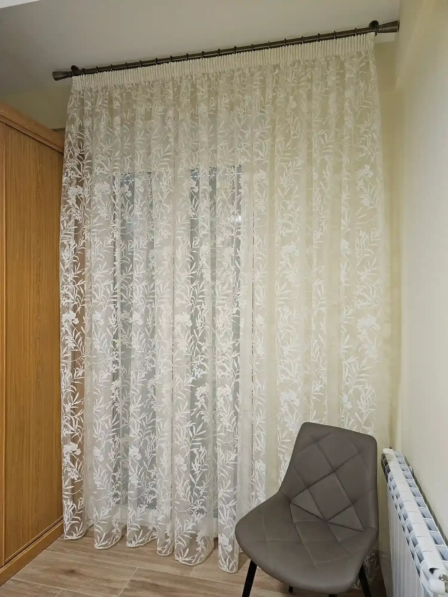Unas cortinas de encaje transparente cuelgan junto a una ventana, al lado de una silla gris y un armario de madera.
