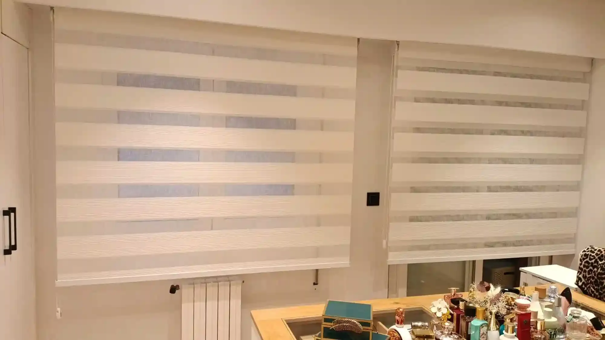 Dos persianas blancas con estampado de cebra cubren las ventanas de una habitación luminosa con una mesa puesta debajo.