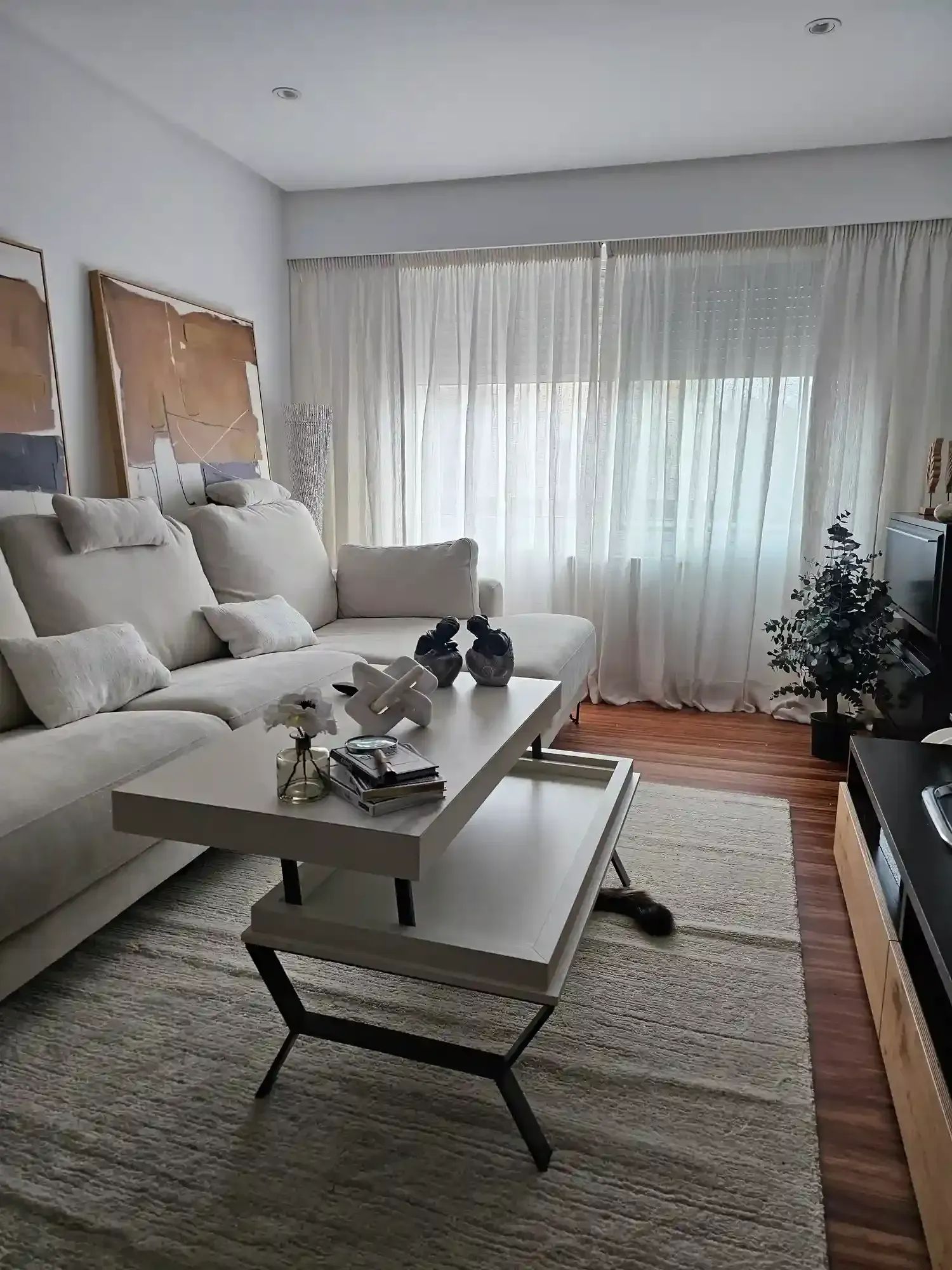 Sala de estar luminosa con sofá blanco, mesa de centro, alfombra y cortinas transparentes en la ventana.