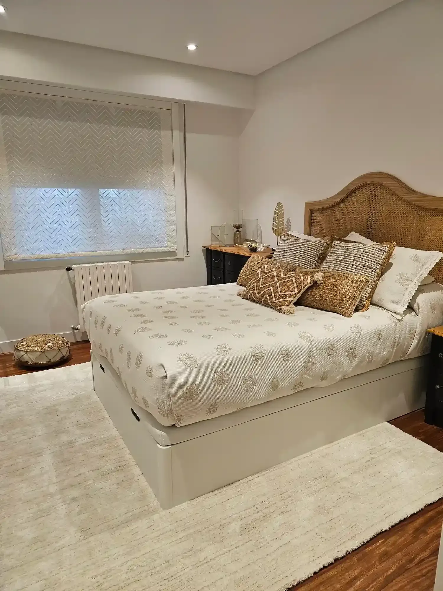 Habitación luminosa con cama blanca, alfombra beige, cabecero tejido y ventana con persianas.