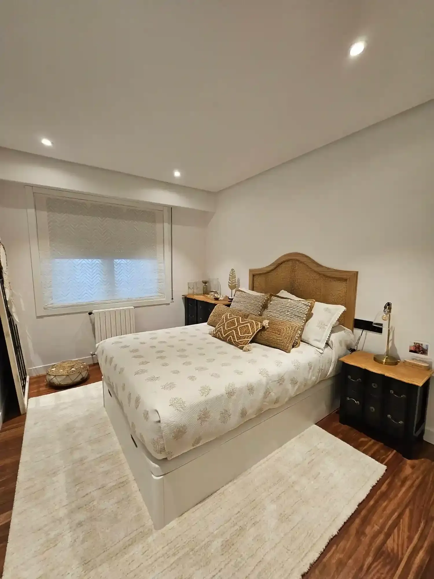 Dormitorio moderno y luminoso con cama blanca, cojines estampados, cabecero de madera y alfombras suaves.