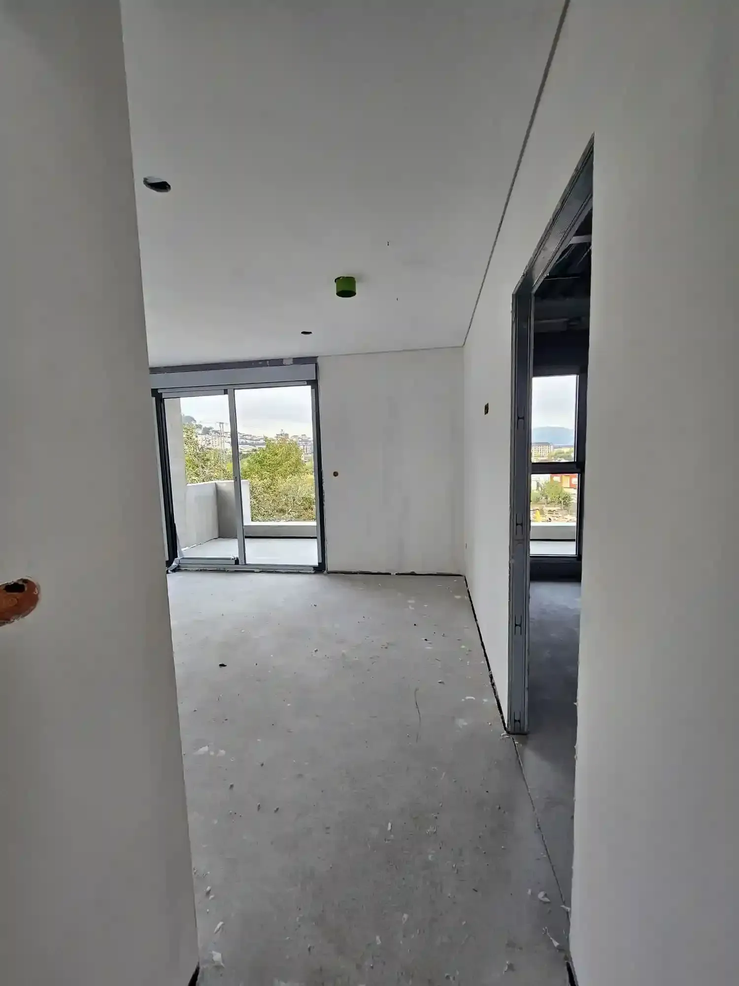 Interior de apartamento vacío y sin terminar con paredes blancas, suelo gris y puertas de balcón.