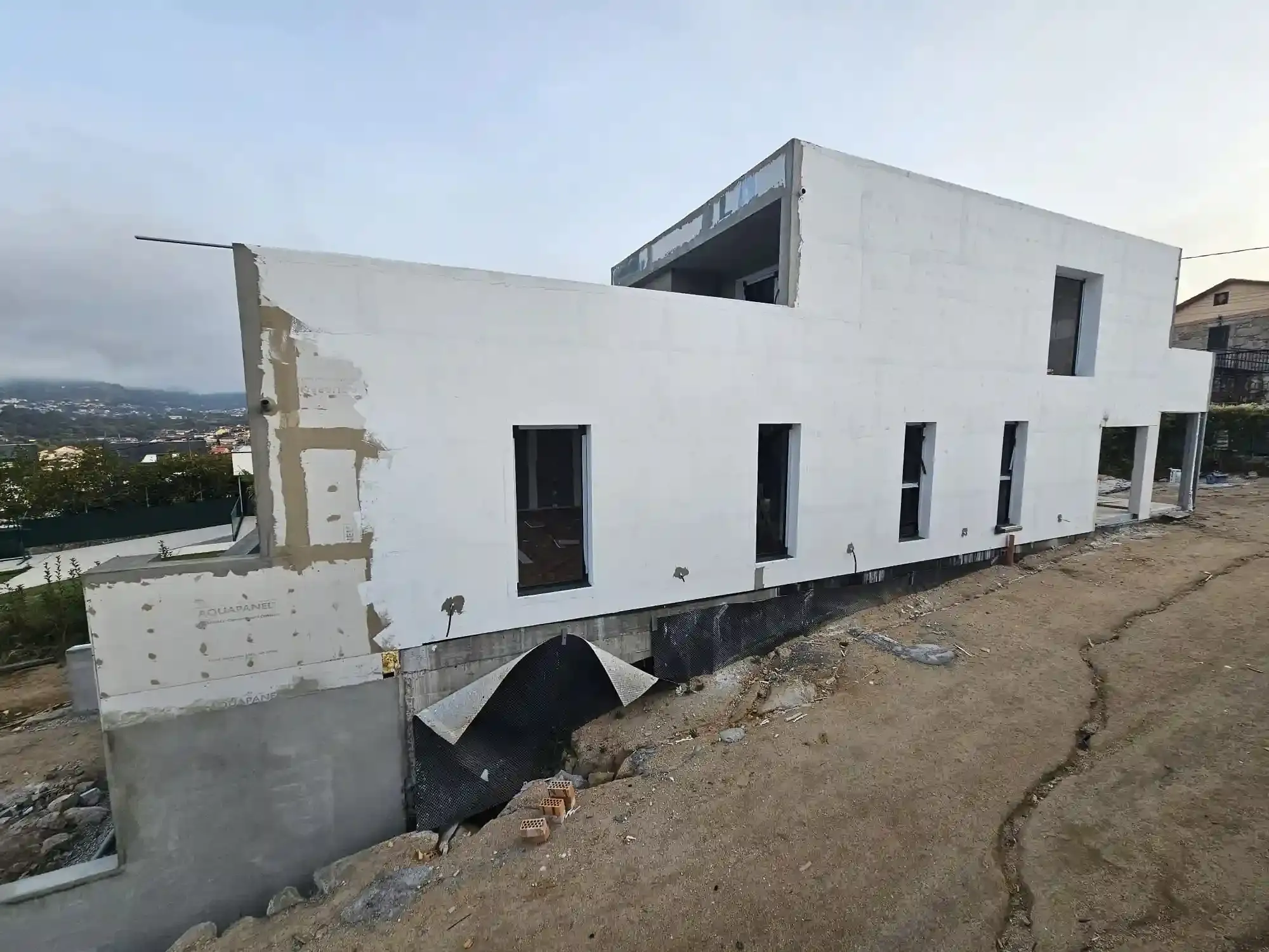 Casa blanca parcialmente construida en una ladera inclinada con cimientos expuestos y paredes sin terminar.