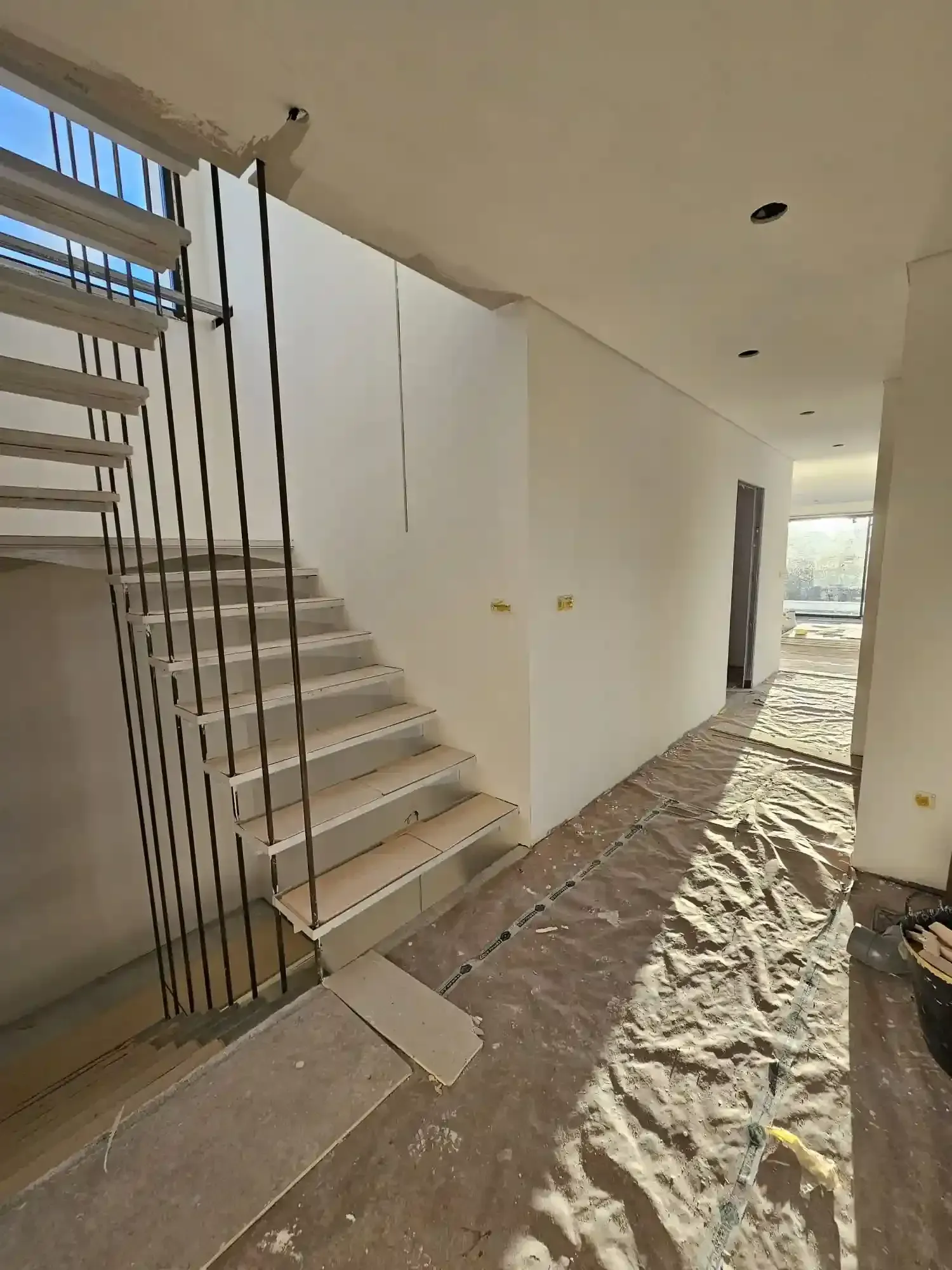 Escalera interior en construcción con escalones a la vista, paredes blancas y luz solar sobre un suelo arenoso.