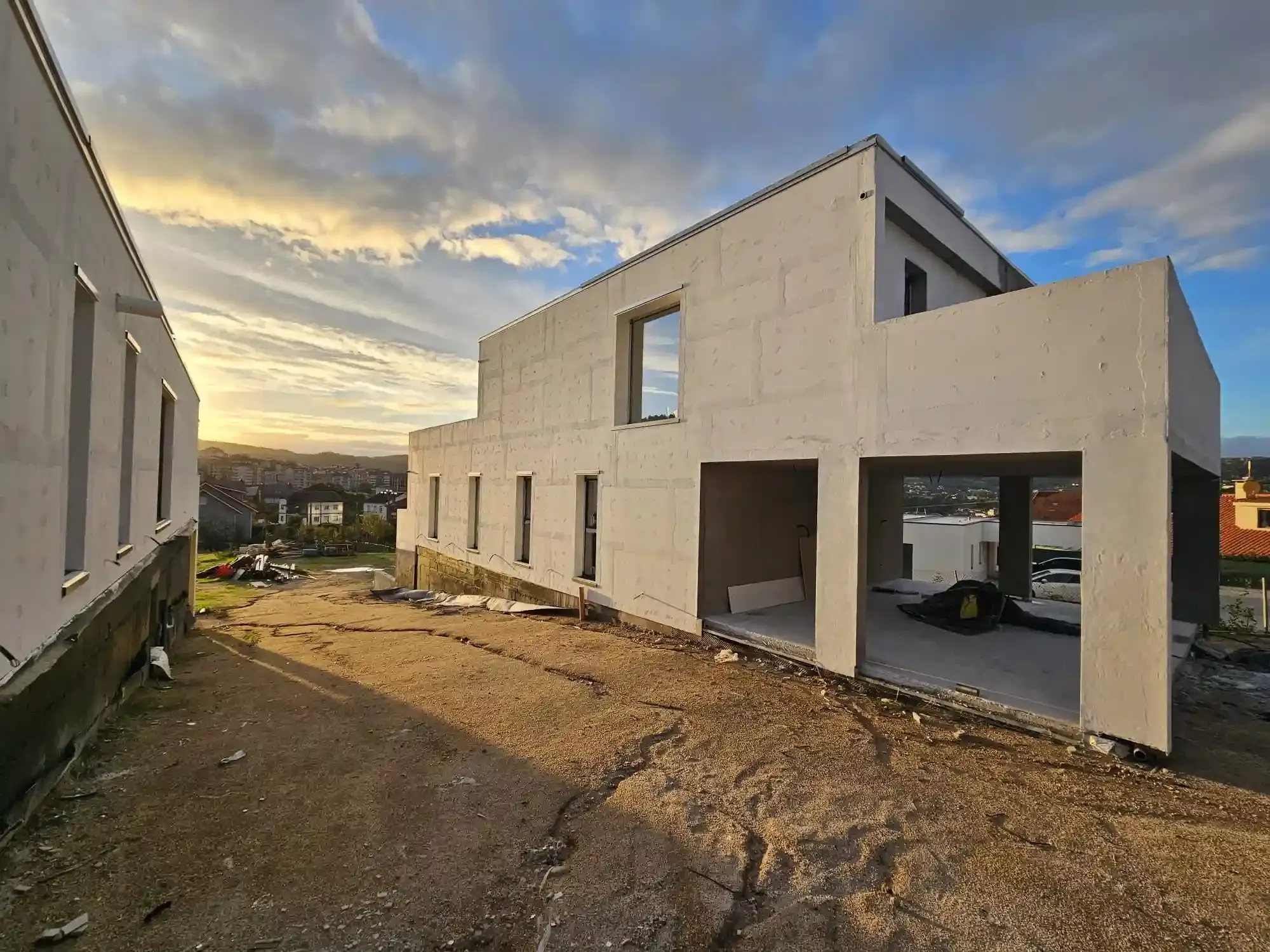 Casa blanca sin terminar con garaje abierto en un terreno de tierra al atardecer, con otro edificio cerca.