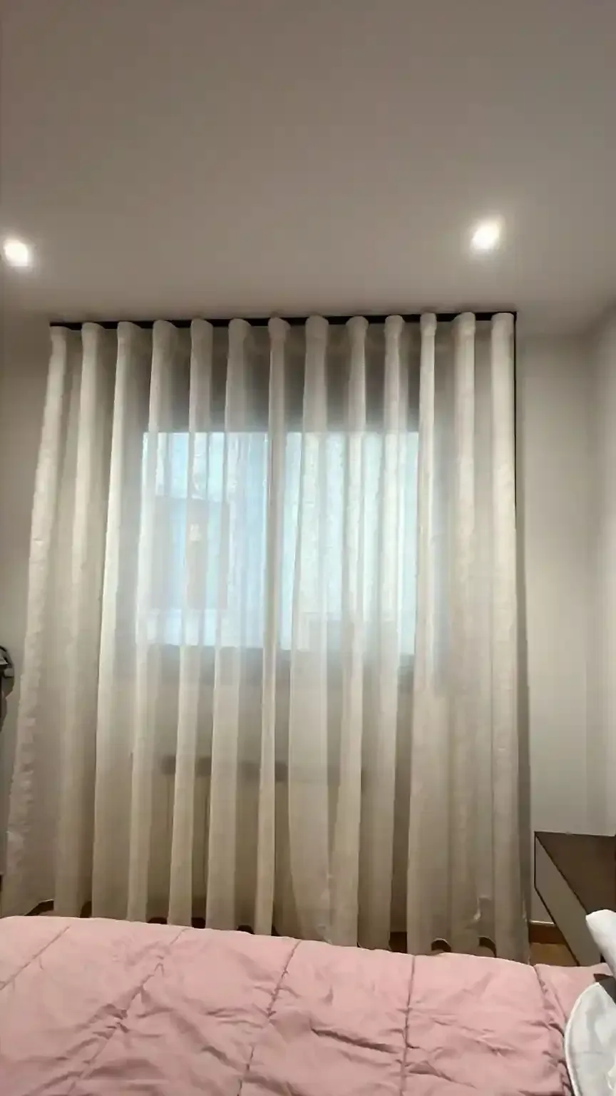 Dormitorio con colcha rosa, cortinas transparentes en la ventana y luces empotradas en el techo.