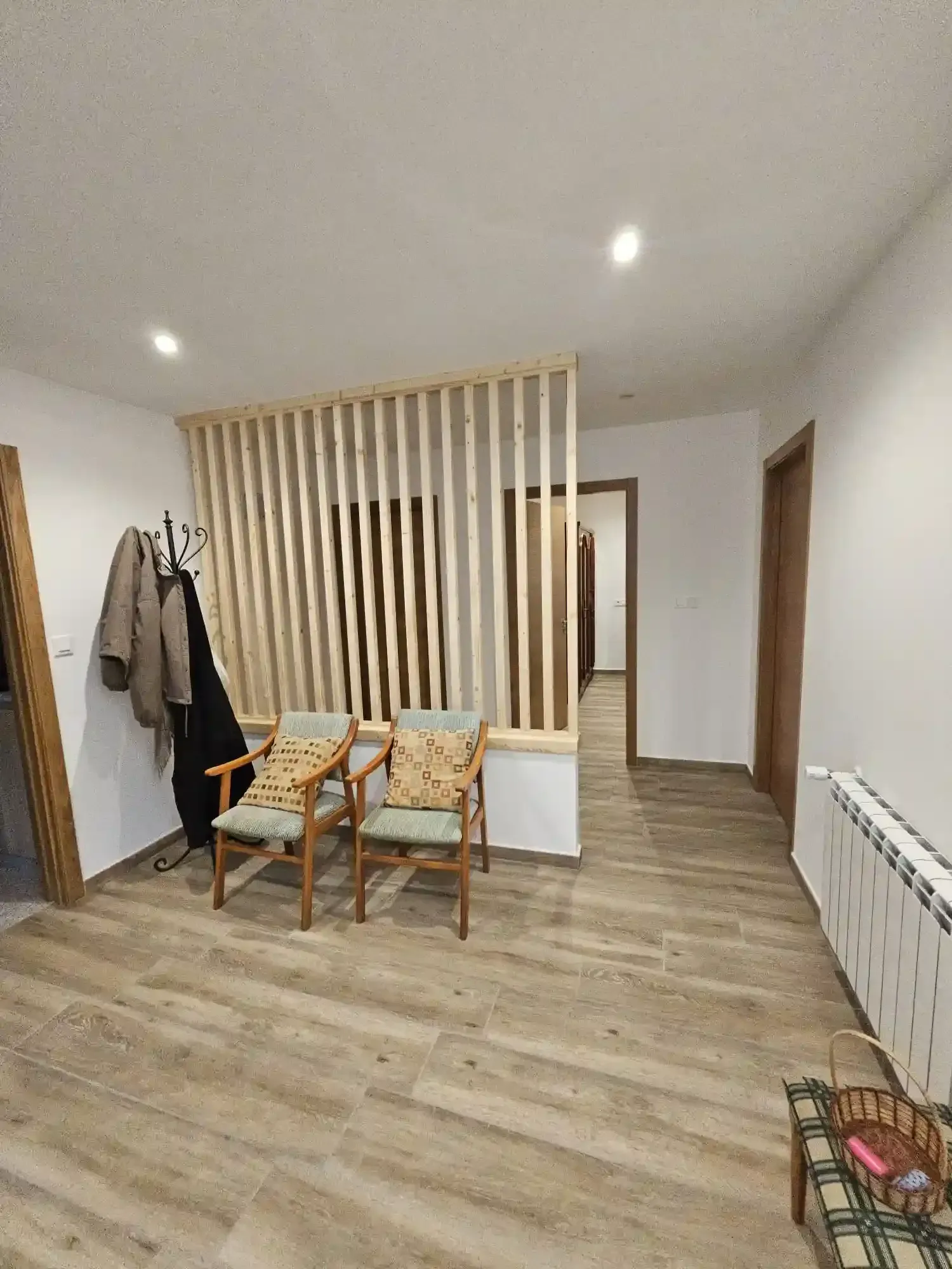 Habitación luminosa y minimalista con suelo de madera, sillas, perchero, separador de ambientes y radiador blanco.