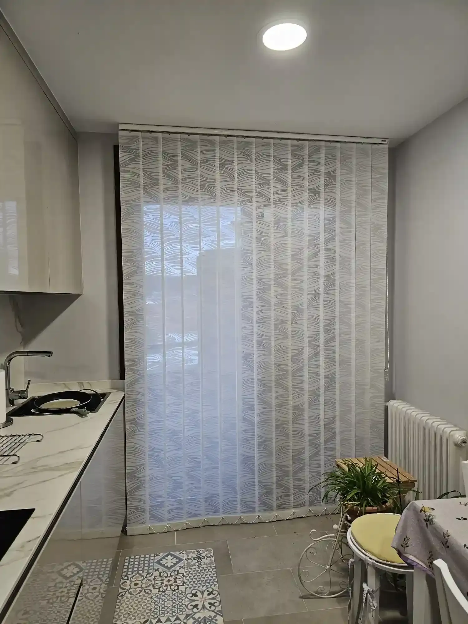 Pequeña cocina moderna con cortinas de encaje, encimera, fogones, radiador y una planta cerca de la ventana.