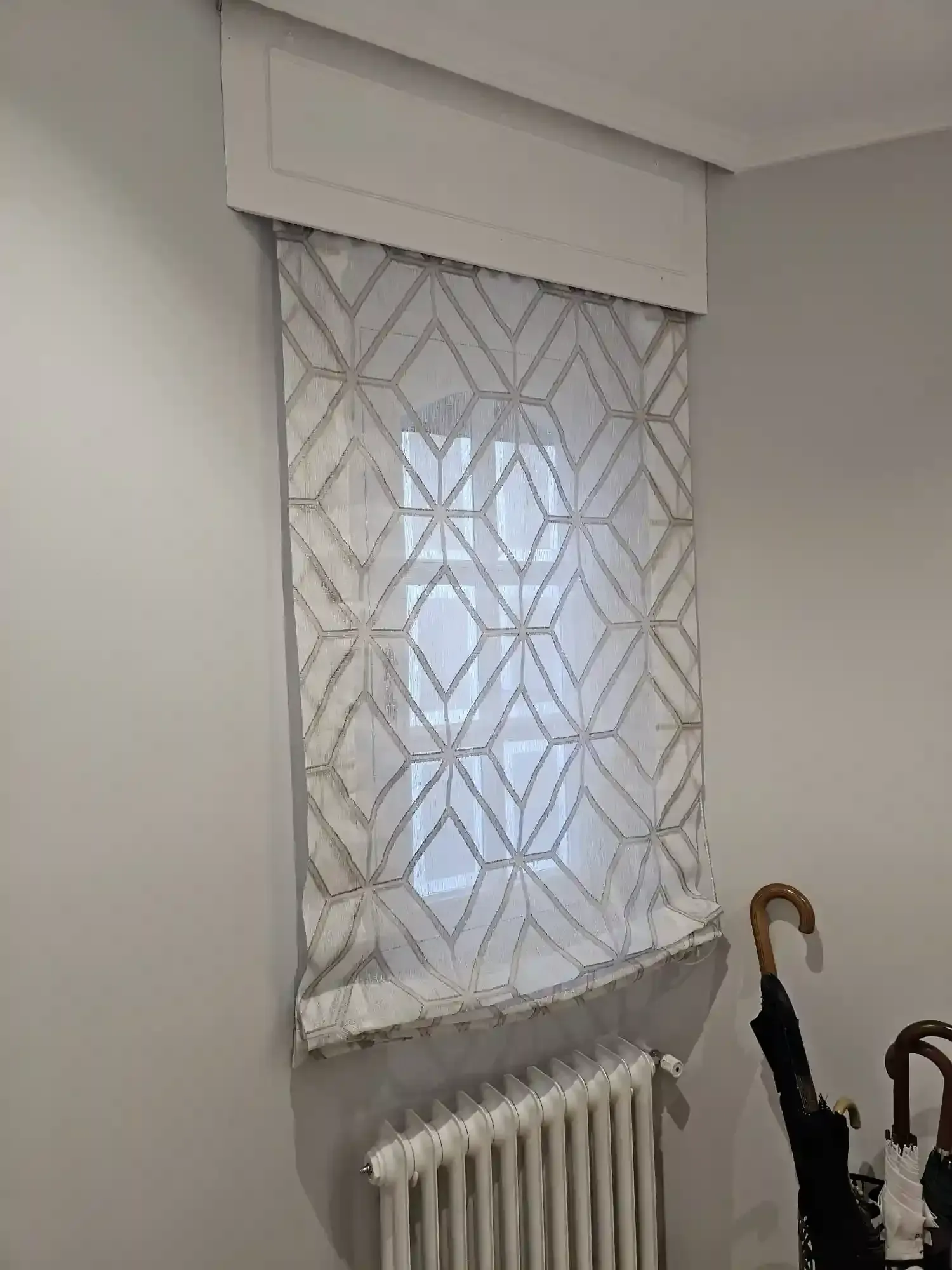 Ventana con cortina blanca estampada y radiador debajo, con un paraguas apoyado cerca.