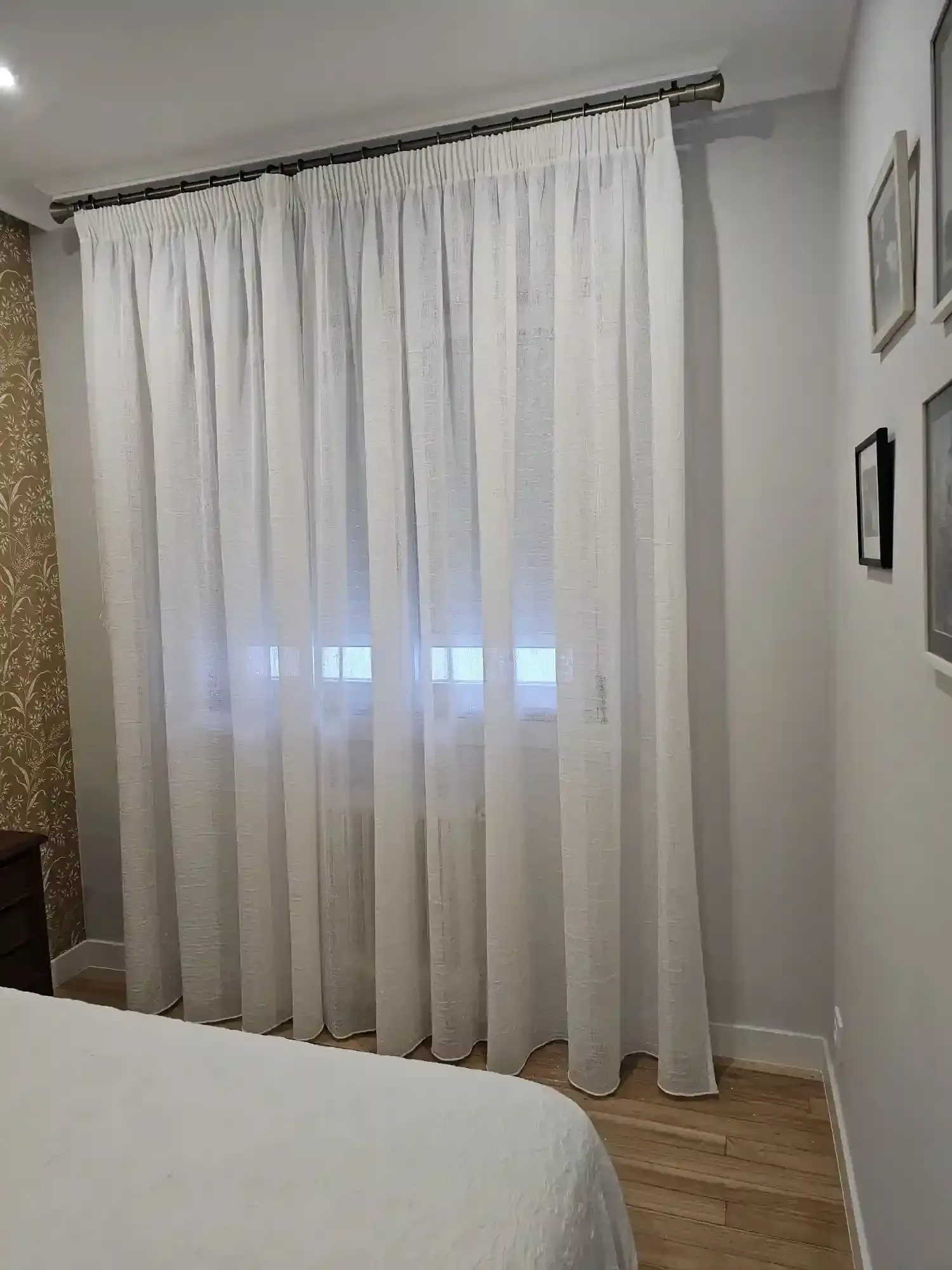 Dormitorio con cortinas blancas transparentes sobre una ventana, suelo de madera clara y parte de una cama en primer plano.