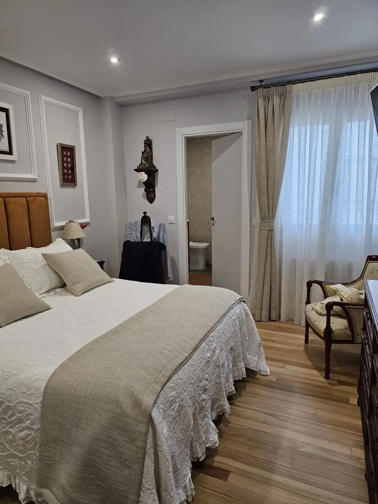 Dormitorio con cama, decoración neutra, suelo de madera y cortinas sobre una ventana luminosa.