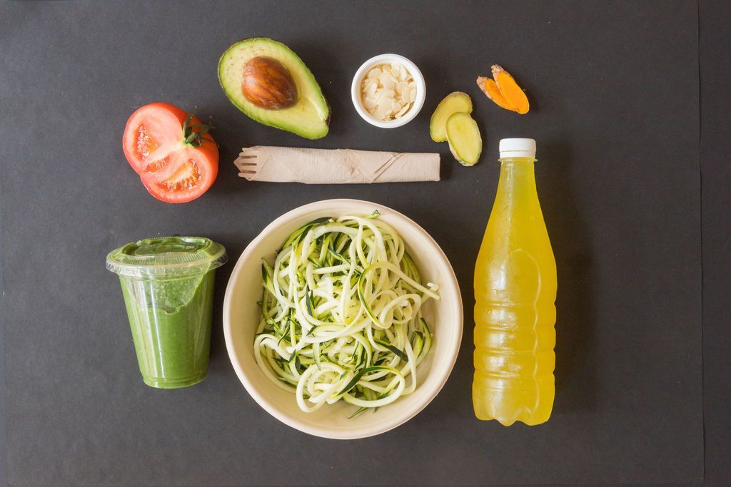 Un tazón de fideos de calabacín rodeado de verduras y una botella de jugo.