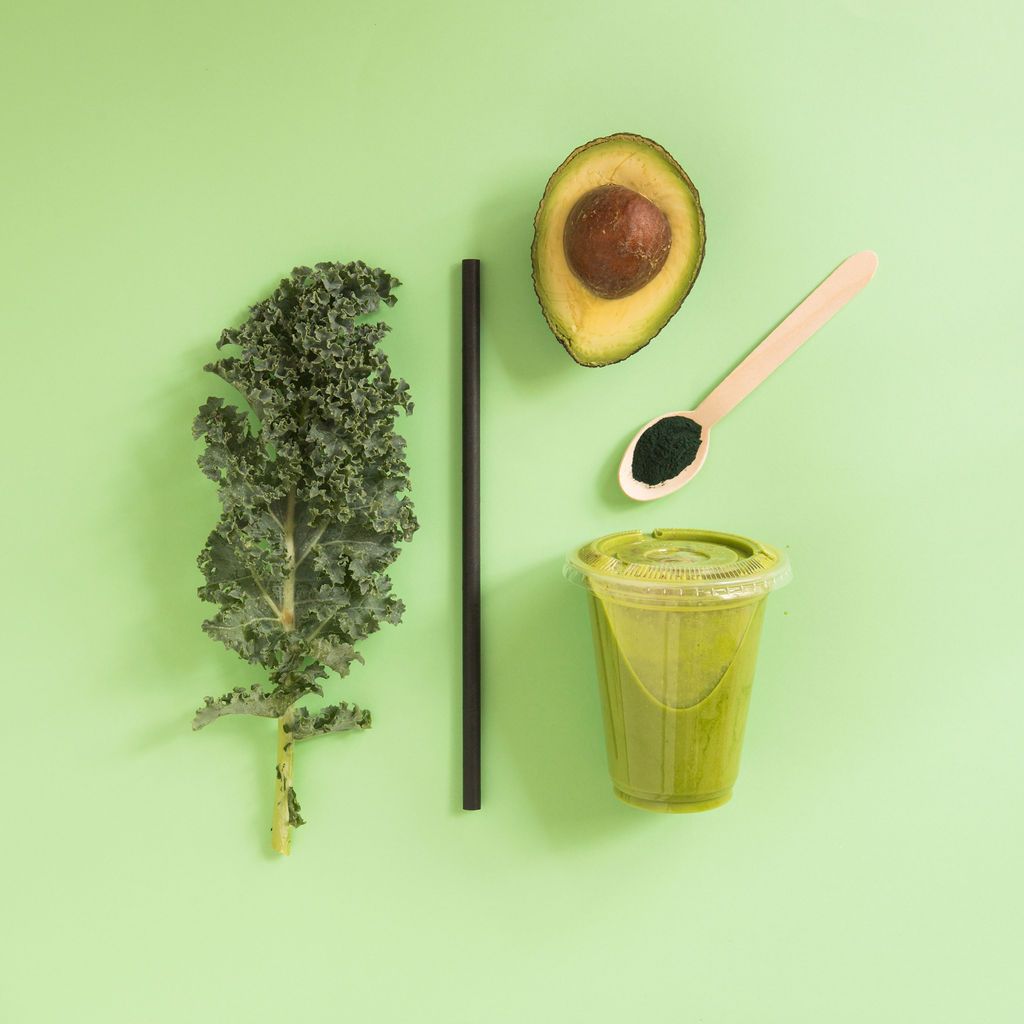 Un batido verde con aguacate, col rizada, una cuchara y una pajita sobre un fondo verde.