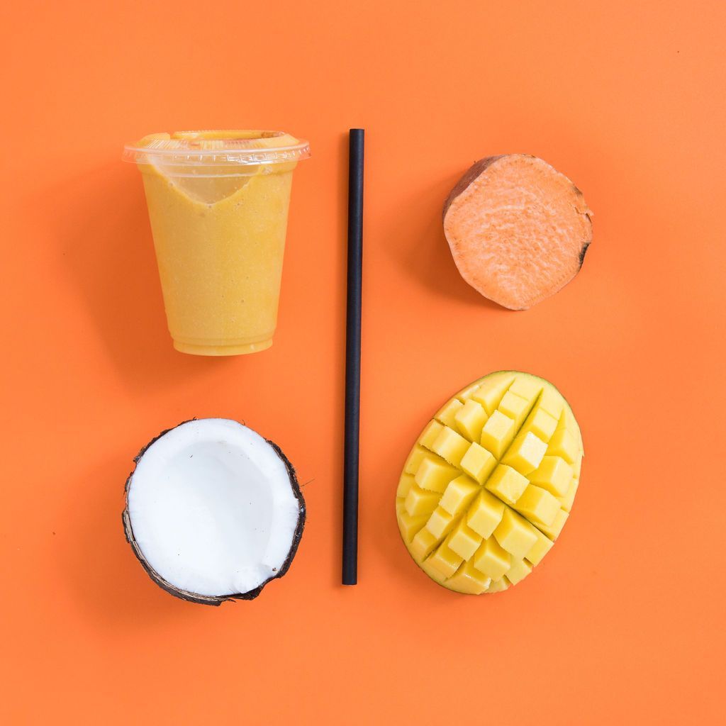 Una taza de jugo, un coco, un mango y una batata sobre un fondo naranja.