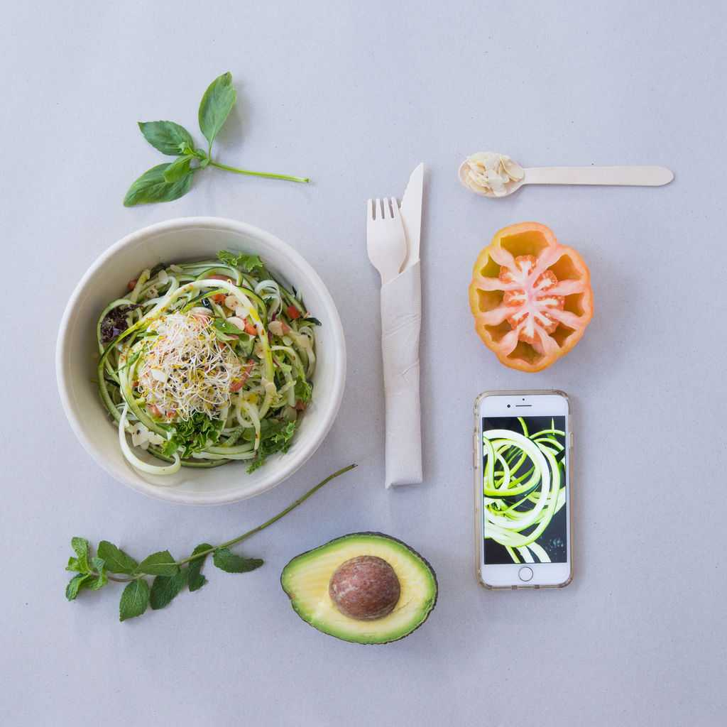 Un tazón de fideos de calabacín está rodeado de verduras y un teléfono celular.