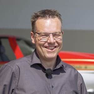 Ein Mann mit Brille und Mikrofon lächelt vor einem roten Auto.