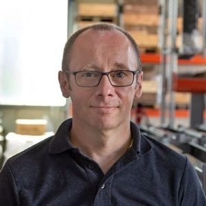 Ein Mann mit Brille und schwarzem Hemd sitzt in einer Lagerhalle.
