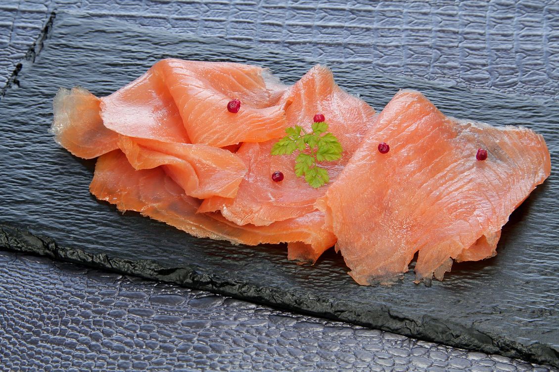 Sashimi de saumon tranché sur une assiette en ardoise sombre, garni d'herbes vertes et de grains de poivre rouge.