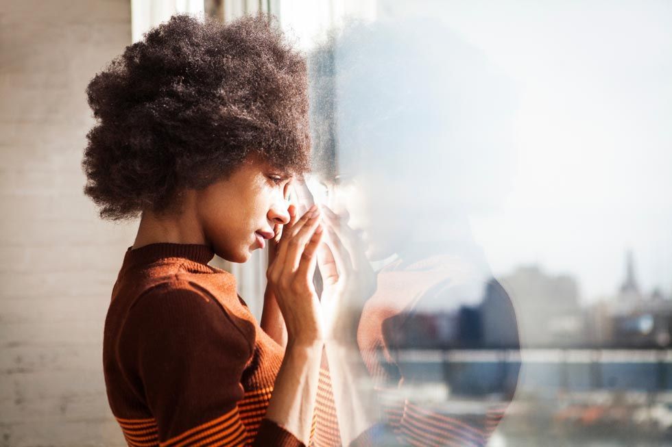 Une femme avec une grande coupe afro, appuyée contre une fenêtre, apparaît triste, reflet du paysage urbain.