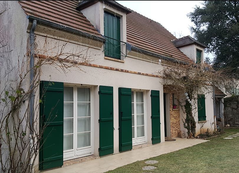 Maison de campagne à la façade en stuc beige, aux volets verts et au toit de tuiles rouges.