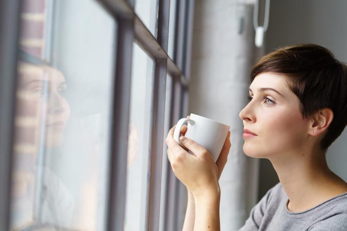 Une femme tenant une tasse blanche regarde par la fenêtre. Son reflet est visible dans la vitre.