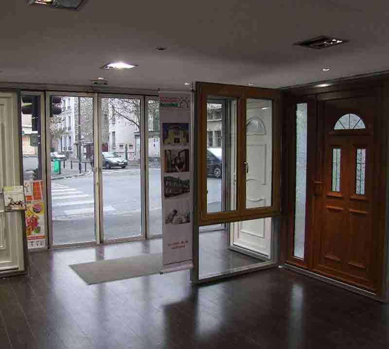 Salle d'exposition avec différentes portes exposées, ouverte sur une vue sur la rue, plancher de bois franc.