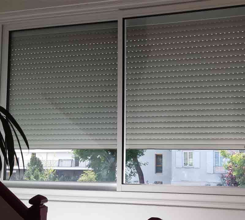 Deux fenêtres avec volets roulants fermés de couleur claire, donnant sur un paysage urbain.