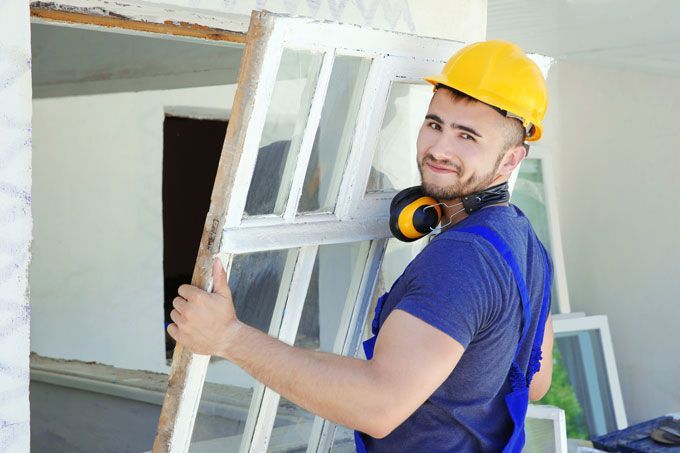 Ouvrier du bâtiment portant une fenêtre, portant un casque jaune et souriant.