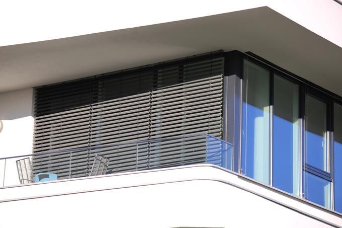 Balcon avec stores horizontaux, fenêtres en verre et garde-corps en métal sur un immeuble moderne.