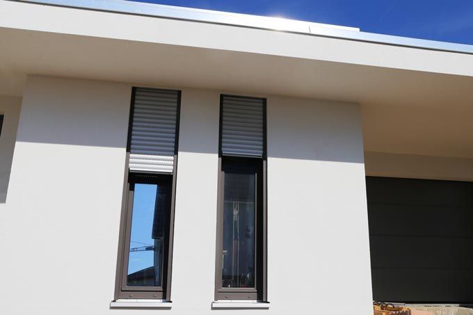 Deux hautes fenêtres avec des stores gris partiellement abaissés sur un bâtiment blanc, sous un ciel bleu.